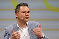 Porträtfoto eines Mannes mit kurzen schwarzen Haaren, der frontal zum Betrachter sitzt und leicht nach rechts blickt. Seine Hände sind leicht unscharf vor dem Körper zu sehen, wie er sie in einer Geste parallel nach oben führt.