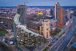 Farbfotografie in der Obersicht von Wolkenkratzern. Das linke halbrunde Gebäude ist treppenartig aufgebaut. Im Hintergrund ist die Stadt zu sehen.