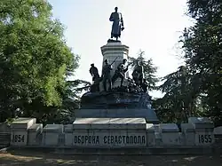 Frontale Farbfotografie eines Denkmals mit kyrillischem Titel und den Jahreszahlen 1854 und 1855 auf einer Steinmauer. Der untere Sockel hat einen gegossenen Felsen mit einigen Soldatenfiguren darauf, die einen weiteren Sockel mit einem General umkreisen. An beiden Seiten des Denkmals sind Bäume.