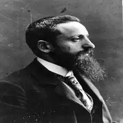 Älterer Mann mit Bart im Seitenprofil nach rechts schauend im dunklen Blazer, weißem Hemd und dunkler Krawatte in schwarzweiß.