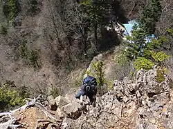 Hier wird eine Person gezeigt, die sich an in den Berg angebrachten Ketten, klettern fortbewegt. Der Hintergrund ist bewaldet.