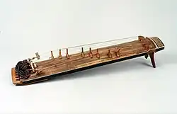 Ajaeng in Spielposition mit Holzbogen, dahinter mehrere Instrumente aufgestellt