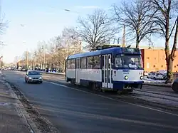 Seitliche Farbfotografie einer blau-weißen Straßenbahn, die über eine Kurve in eine Allee mit alten Gebäuden fährt. Rechts hinten ist ein weißes Gebäude mit rundem Mittelteil und grünem Dach sowie Ornamenten.