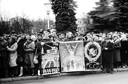Frontale Schwarzweißfotografie von einer Menschenmenge an einem Bürgersteig. In der Mitte werden drei Plakate mit kyrillischer Schrift „CCCP“ gehalten, was die ehemalige Sowjetunion darstellt. Rechts neben den Plakaten steht ein alter Mann mit Fotoapparat.
