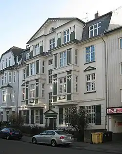 Wohngebäude mit einer aufwendigen Fassade. Das Gebäude hat mehrere Etagen und ist in den Farben Weiß und Grün gestaltet. Die Architektur weist dekorative Elemente auf, darunter Pilaster, Gesimse und Fensterrahmen mit filigranen Designs. Es gibt mehrere Fenster auf jeder Etage, einige davon mit Fensterläden. Der Eingangsbereich wird von einem kleinen Portikus mit einem dreieckigen Giebel betont. Vor dem Gebäude stehen zwei geparkte Autos, und es sind Büsche sichtbar, die möglicherweise Teil eines Gartens sind. Auf der rechten Seite des Gebäudes befindet sich ein Schild mit der Aufschrift „LAGER-VERK“ sowie der Zahl „55“.