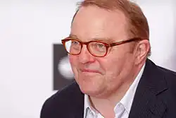 Gustav Peter Wöhler bei der Verleihung des Studio Hamburg Nachwuchspreises 2012 trägt einen dunkel gestreiften Anzug und ein weißes Hemd mit Kragen. Er hat hellbraune Haare und trägt eine braune Brille. Der Hintergrund des Bildes ist weiß und zeigt ein schwarzes, kreisförmiges Muster, das hinter der Person sichtbar ist.