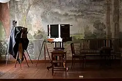 Farbfotografie von drei Balgenkameras, die vor einer Wand mit Landschaftsmalerei stehen. Die Kameras werden von links nach rechts immer größer und stehen auf Stativen. Rechts befindet sich eine Sitzgruppe.