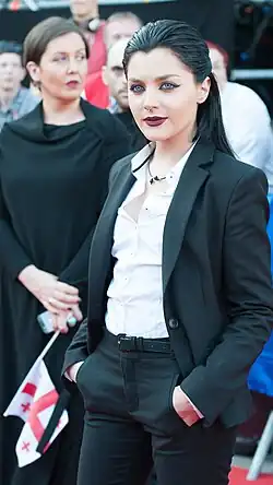 Die junge Sängerin blickt an der Kamera vorbei. Sie hält die Hände in den Taschen ihres schwarzen Hosenanzug. Ihr schwarzes Haar ist mit viel Gel nach hinten gezogen und zu einem Zopf gebunden.