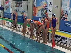 Sechs Niederländerinnen stehen am Beckenrand. Sie tragen eine blaue Bademütze und einen blau-orangenen Badeanzug. Hinter ihnen befestigt die Torhüterin ihre rote Bademütze.