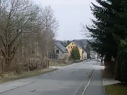 abschüssige Straße ohne Straßenbahnmarkierungen, Bürgersteig auf der rechten Seite, links und rechts Bäume, hinten ein paar Häuser, Autos und Ampel