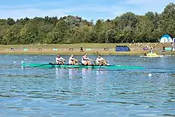 Vier Ruderer sitzen mit weißem Hemd und rotem Brustring im grünen Boot.