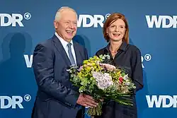 Tom Buhrow überreicht Katrin Vernau einen Blumenstrauß vor blauer Wand mit WDR-Logo, beide schauen zum Publikum