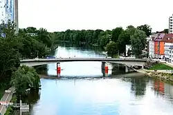 Blick aus erhöhter Position auf Donau und Gänstorbrücke. In der Mitte fließt die Donau nach hinten hin weg. Sie ist auf beiden Seiten von intensiv grün belaubten Bäumen gesäumt. In der Mitte verläuft die Gänstorbrücke waagerecht von links nach rechts. Unter der Brücke ist rechts und links je ein Hilfspfeiler erkennbar, an der Stirnseite ist je eine rote Matte angebracht. Rechts neben der Brücke ist Bebauung auf der Neu-Ulmer Seite sichtbar, links hinter der Brücke ragt das Maritim-Hotel in Ulm in die Höhe.