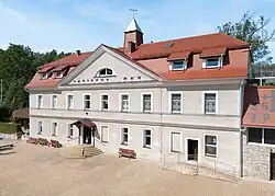 Ein breites Gebäude mit zwei Stockwerken, wobei sich das Zweite im Dach befindet. Es besitzt einen Glockentürmchen in der Mitte des Daches. Es besitzt ein großes Zwerchdach in der Mitte.