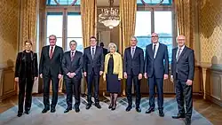 Acht Personen in formeller Kleidung, die in nebeneinander vor zwei großen Fenster in einem prunkvollen Raum stehen.