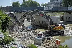 Blick durch einen Bauzaun in eine Baustelle. Im Hintergrund ist eine Brückenfahrbahn von links nach rechts durchs Bild laufend erkennbar. Im Vordergrund steht ein Raupenbagger auf einer Rampe, neben ihm die Reste der abgebrochenen westlichen Brückenhälfte.