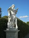 Statue von Melpomene im Park Belvedere, Wien, Schrägansicht von vorn links; Melpomene breitet den rechten Arm in Richtung Betrachter aus, in der linken hält sie eine große Muschelschale; ihr rechtes Bein ist nach vorn gebeugt, so dass das Schuhwerk erkennbar ist, der Fuß steht auf einem kleinen Baumstumpf; rechts am Boden eine übergroße Theatermaske