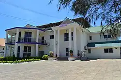 Ein weißes Haus von vorne, mit Balkonen auf der linken Seite und einem von Säulen getragenen Vordach auf der rechten.