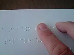 Zeige- und Mittelfinger eines Lesenden gleiten über die obere von zwei Zeilen Braille-Text. Deutlich zu erkennen sind die punktförmigen Erhöhungen in dem weißen Papier, die die Schriftzeichen bilden.