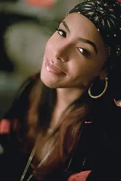 Aaliyah in Berlin (2000), trägt ein schwarzes Kopftuch mit einem floralen Muster und große Creolen-Ohrringe. Ihr langes, gewelltes Haar ist braun an den Spitzen und dunkler an den Ansätzen. Sie ist in einem schwarzen Outfit mit roten Akzenten gekleidet. Der Hintergrund ist unscharf, wodurch keine weiteren Details erkennbar sind.
