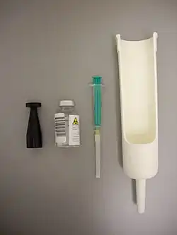 Von links nach rechts: Prüfstrahler, Glas­flasche für Radionuklid in wässriger Lösung, Spritze und Proben-Einsatz.