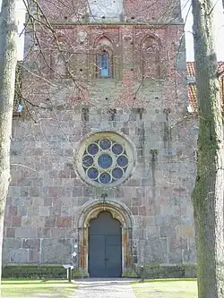 Westfassade: Sand­stein­portel (Rund­stäbe, Blatt­kapitelle), Rosen­fenster, Fenster­etage (Rund­stäbe, Back­stein teilw. glasiert)