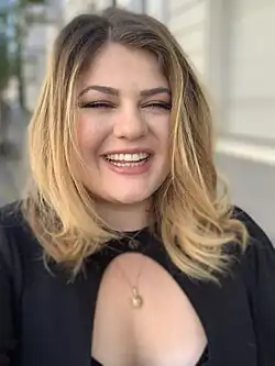 Alina (2019) mit schulterlangen, welligen, blonden Haaren, die ein schwarzes Oberteil mit einem Cut-Out-Design im Brustbereich trägt. Sie lächelt und trägt zwei Halsketten – eine mit einem kreisförmigen Anhänger und die andere mit einem sonnenförmigen Anhänger. Der Hintergrund wirkt wie eine Außenszene, mit verschwommenen Gebäuden und grünen Flächen im Blick.