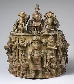 Zylindrisch geformte Gelbguss-Skulptur von 33 cm Höhe und knapp 36 cm Breite und Tiefe mit einem Fries. In der Mitte des Fries steht der Ezomo Ehenua in vollem militärischen Ornat und Kriegstrophäen umklammernd umringt von einer Gruppe kleiner dargestellter Soldaten, Diener und Priester. Auf dem Zylinder befinden sich zwei Reihen von Musketieren, von denen ein Teil portugiesische Soldaten sind. Vor diesen Musketieren befindet sich eine kleine Skulpturengruppe mit Oba Akenzua I. Der Oba bringt seinen Ahnen Opfer dar, damit der Ezomo siegen und über seine Widersacher triumphieren kann.