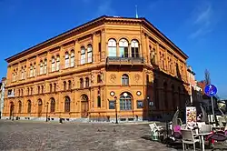 Eckansicht in Farbe von einem braunen dreigeschossigen Gebäude im Renaissance-Stil, das Säulen und Figuren zwischen den Fenstern hat. Die Rundbogenfenster sind mit Sprossen verziert. Eine mittlere Fassade ist zwischen den beiden längeren Fassaden quer angebaut. An dieser Fassade hängen im Erdgeschoss drei Uhren und im Obergeschoss ist ein kleiner Balkon zu sehen. Auf dem Dach ist eine Turmhaube mit zwei Uhren.