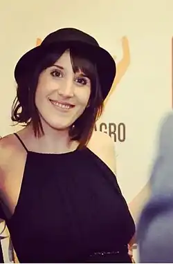 Laia Alvarez mit schulterlangen, dunklen Haaren, die ein ärmelloses schwarzes Kleid und einen schwarzen Hut trägt. Im Hintergrund ist eine helle Wand zu sehen, auf der teilweise der Schriftzug „AGRO“ zu erkennen ist. Sie steht mit leicht angehobenen Armen. Auf der rechten Seite des Bildes ist eine weitere Person teilweise sichtbar, die ein helles Kleidungsstück trägt.