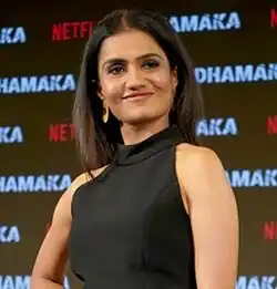 Amruta Subhash bei der Filmpremiere von Dhamaka 2021. Der Hintergrund ist dunkel gehalten und mehrfach das Wort „DHAMAKA“ in weißer und roter Schrift sowie das Netflix-Logo abbildet. Amruta Subhash trägt ein ärmelloses schwarzes Kleid, das elegant und schlicht wirkt. Sie hat glattes, schulterlanges Haar und steht in einer selbstbewussten Pose, die den Eindruck von Stärke und Stil vermittelt.