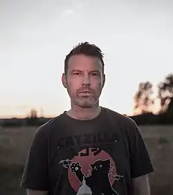 Halbprofil eines lächelnden blonden Mannes mit kurzen Haaren und Dreitagebart vor einem weiten Feld