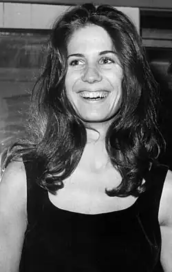 Schwarz-weiß Foto mit der lächelnde Anne Byrne Hoffman mit langen, dunklen Haaren, die ein ärmelloses, dunkel gefärbtes Oberteil trägt. Der Hintergrund scheint sich drinnen zu befinden, möglicherweise in einem Raum mit einem Fenster oder Spiegel.