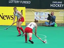 Die beiden Engländerinnen tragen ein weißes Trikot und einen roten Rock. Richardson schlägt den Ball von außerhalb des Schusskreises, Panter wartet im Schusskreis auf den Pass.