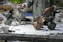 Auf einer Werkbank stehen mehrer Versionen eines Kopfes einer antiken Frauenstatue – einmal mit rötlicher Färbung, einmal schwarz und zwei graue Versionen. Dahinter der Oberkörper eines Mannes in einem grauen Pulli, der gerade eine der grauen Köpfe mit der rechten Hand berührt.