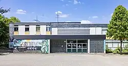 Zentraler Blick auf ein 60er-Jahre-Schulgebäude mit drei flachen, nach hinten höher werdenden Bauteilen. Der vordere, flache Eingangsbereich mit einem Wandgemälde und dem Schriftzug „Archiv“