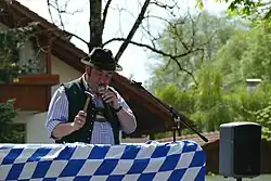 Ein Mann mit traditioneller bayerischer Kleidung steht an einem mit weiß-blauen Ruten verzierten Pult, in einer Hand hält er ein Mikrofon, in der anderen einen erhobenen Hammer