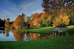 Farbfotografie eines Sees mit einer kleinen Bogenbrücke aus Stein am rechten Bildrand. Im Hintergrund eine Wiese und Herbstbäume. Im Vordergrund ist ebenfalls eine Wiese.
