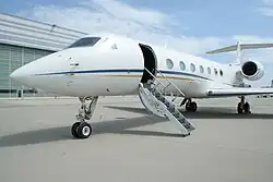 Seitenansicht mit offener Tür eines Privatjets des Flugzeugherstellers Gulfstream, Modell G550, die Tür ist offen, die Treppe ausgeklappt, man sieht sieben liegende ovale Fenster (die typische Fensterform von Gulfstream), das Flugzeug steht auf dem Vorfeld eines Flughafens vor einem Hangar bei strahlend blauem Himmel