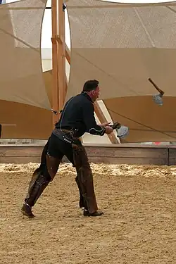 Man sieht einen nach recht blickenden, stehenden Mann in einem Westernkostüm von der Seite, der ein Bein weiter vorne hat als das andere. Vor ihm in der Luft eine fliegende Axt, zwei weitere Äxte hält er in der rechten Hand..