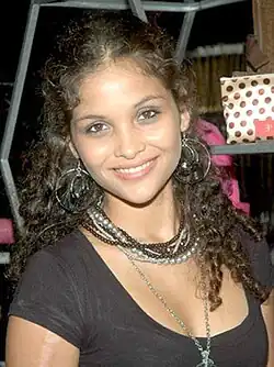 Lächelende Ayesha kapur (2012) mit lockigem Haar, die ein schwarzes Oberteil und mehrere Halsketten trägt. Im Hintergrund befindet sich ein Regal mit verschiedenen Gegenständen, darunter eine Tasche mit Tupfenmuster.