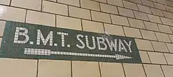 Cremefarben in Halbverband geflieste Bahnhofswand mit einem Wegweiser aus einem braunen Keramik-Mosaik mit der Inschrift „B.M.T. Subway“ und einem Richtungspfeil.