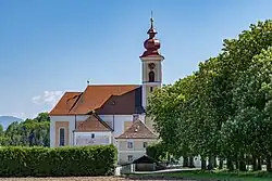 Fotografie der Kirche bei Sonnenschein und blauem Himmel. Im Vordergrund rechts ist eine auf die Kirche zuführende Kastanienallee, links im Vordergrund eine hohe Hecke. Im Hintergrund hinter der Kirche sind einige hohe Bäume zu sehen.