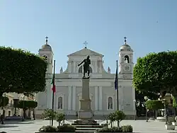Das Bild zeigt eine weiße Kirche, vor der eine schwarze Soldatenstatue sowie eine Italien- und eine EU-Flagge steht.