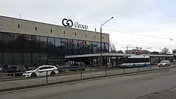 Seitliche Farbfotografie von einem Gebäude mit großer Glasfront und Reklame an dessen rechter Seite. Im oberen Bereich ist eine helle Steinfassade und auf der rechten Dachseite stehen große Reklamebuchstaben. Vom Eingang geht bis zum rechten Bildrand eine Überdachung, vor der ein blau-weißer Bus steht. Links parken einige Autos und zwei Autos fahren im Vordergrund auf der Straße.