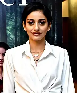 Banita Sandhu mit dunklem Haar, die ein weißes Hemd mit einem Kragen trägt und hat einen neutralen Gesichtsausdruck. Das Hemd ist bis oben zugeknöpft und hat am unteren Ende einen Knoten, der das Kleidungsstück interessant gestaltet. Sie trägt außerdem zwei Halsketten: Die eine hat zwei runde Anhänger, und die andere ist eine schlichte Kette. Im Hintergrund befindet sich ein Poster mit einer Waldszene sowie einer teilweise sichtbaren weiteren Person.