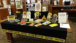 Tisch in einer Bibliothek mit einer Auslage von Büchern; auf einem Schild steht: „Stand up for your right to read – read a banned book“; an und zwischen den Büchern sind gelbe Gefahren-Warnschilder angebracht mit der Aufschrift: „CAUTION“ bzw. „Caution – Banned Books Area“
