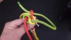 Nun nimmt man das grüne Kardeel und zieht es entgegen der Uhrzeigerrichtung unter dem roten Auge durch. Das grüne Kardeel liegt parallel zum gelben Kardeel