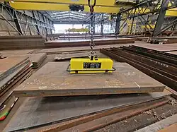 Ein elektropermanenter Lasthebemagnet EPMB 5000 hebt eine schwere Stahlplatte in einer großen Lagerhalle. Der Magnet ist am Kranhaken befestigt und verfügt über eine Fernsteuerung.