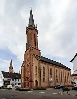 Blick von der Hauptstraße auf das Kirchengebäude, mit dem Turm der katholischen Pfarrkirche im Hintergrund